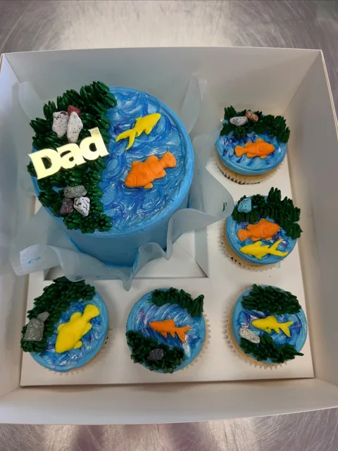 Father's Day fish pond cake with blue buttercream water, piped green reeds, chocolate rocks, orange and yellow fondant fish and a gold Dad topper, boxed with five matching cupcakes