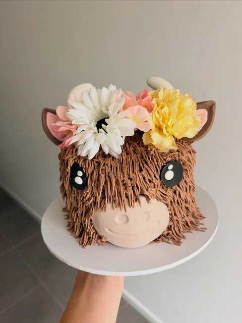 Highland cow cake with shaggy piped brown buttercream fur, a fondant face and horns, and a crown of buttercream flowers