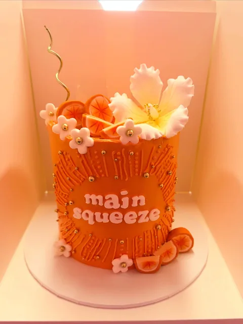 Tall orange cake with textured piping, main squeeze written on the front, orange slices and white sugar flowers