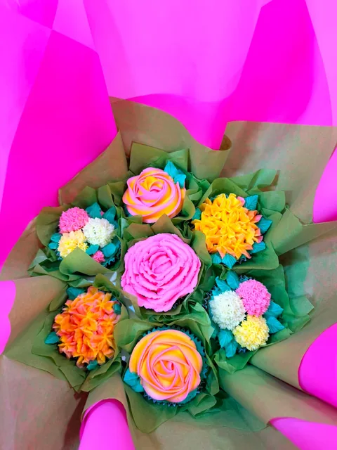 Cupcake bouquet of piped buttercream roses, chrysanthemums and pompom flowers in pink, orange, yellow and teal, wrapped in green and kraft paper