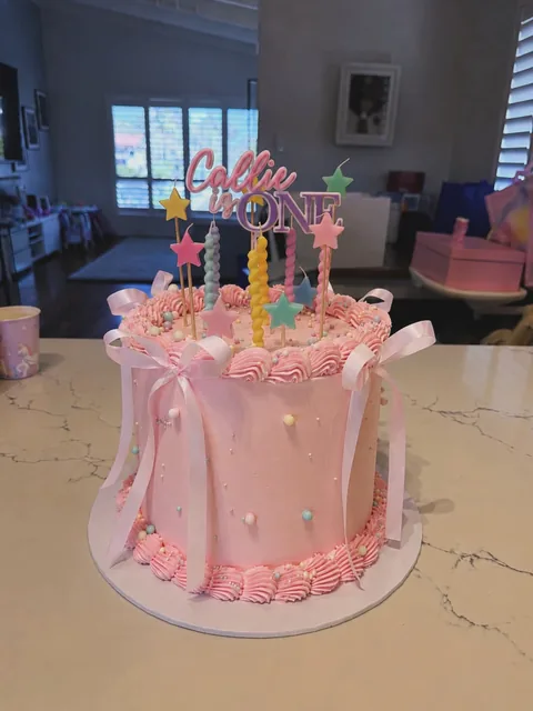 Tall pale pink cake with ribbon bows, pearls, pink piping, star candles and a personalised ONE topper for a first birthday