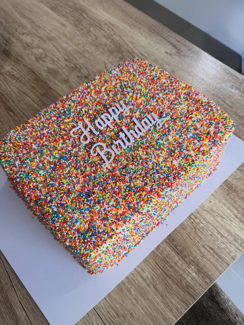 Rectangular slab cake completely covered in rainbow hundreds and thousands with a white Happy Birthday script topper