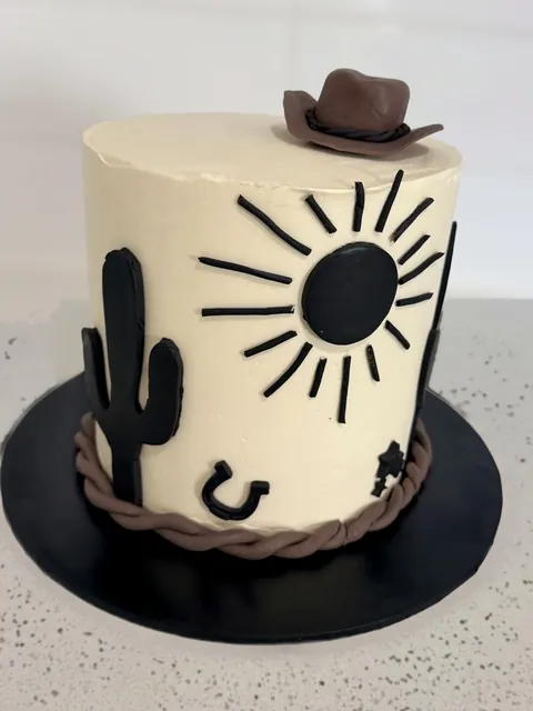 Cream cowboy-themed cake with black cactus, sun and horseshoe and a brown cowboy hat on top